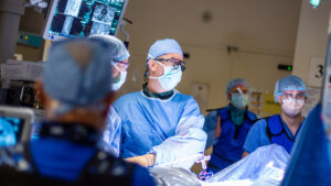 A surgeon looks toward a monitor while performing a laparoscopic procedure on a patient. Other surgical team members are visible around the room.
