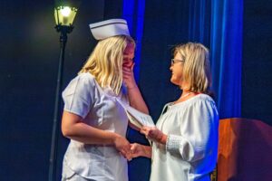 A woman dressed in a white nursing uniform presses her left hand against her face, sobbing, as she accepts a certificate and a pin from an instructor.