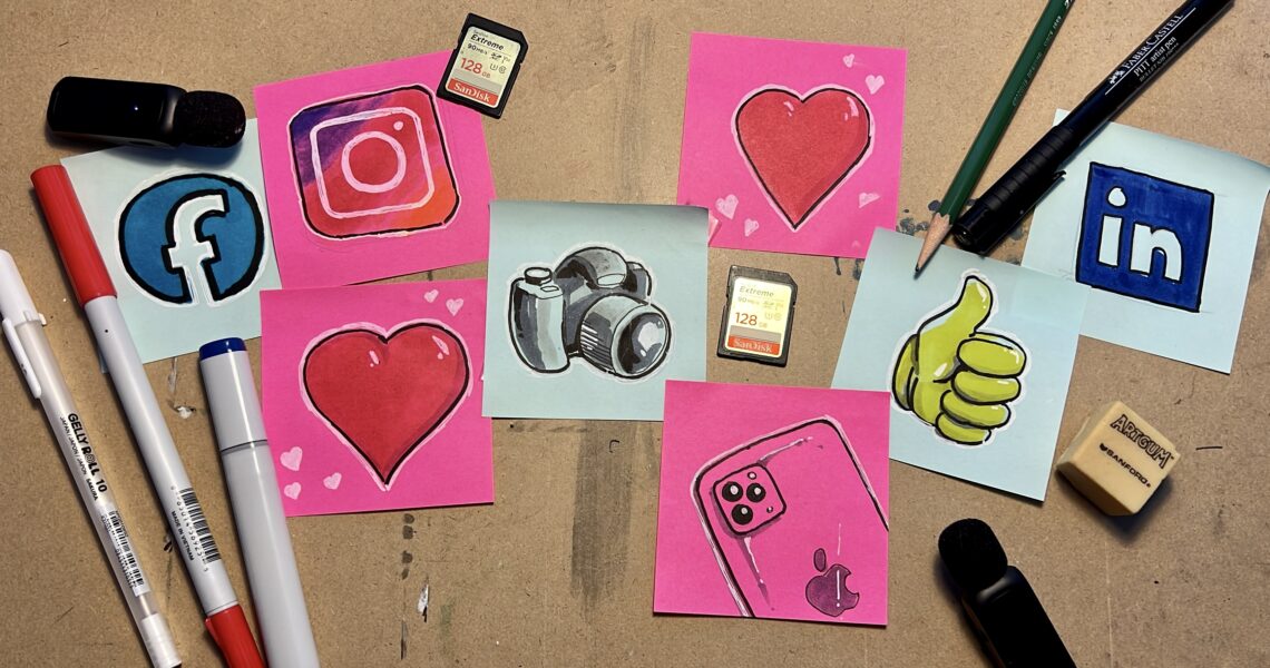 Several pink and blue sticky notes, each containing an illustrated icon of social media logos, emojis, an iphone and a camera, sit on a drawing table. They are surrounded by pencils, pens, memory cards, microphones, and an eraser.